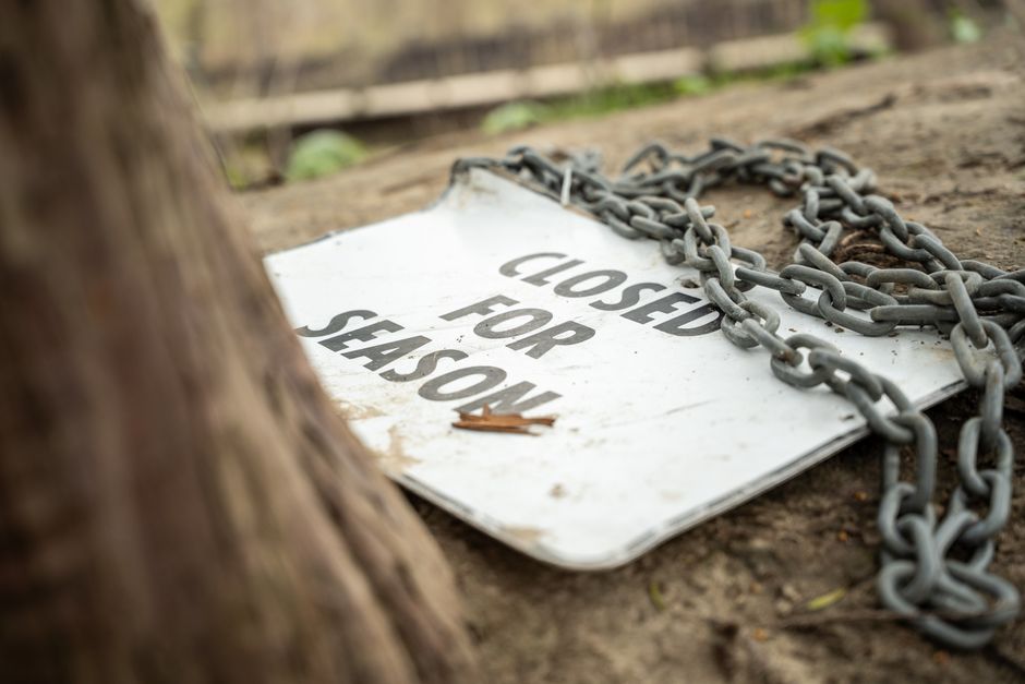 A wooden gate with a warning sign attached, situated in a rural outdoor setting with lush green grass, flowerbeds, and large trees in the background under a partly cloudy sky. The sign reads, 'WARNING