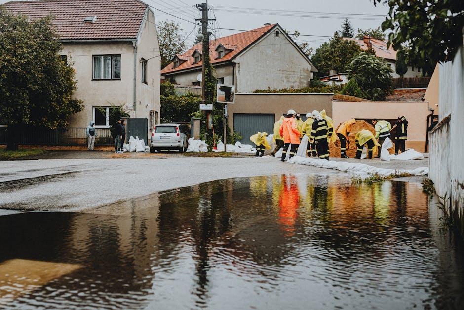 A group of workers in high-visibility safety jackets and protective gear are conducting flood clean-up operations in a residential area, visible with water pooling on the street surface. The scene inc