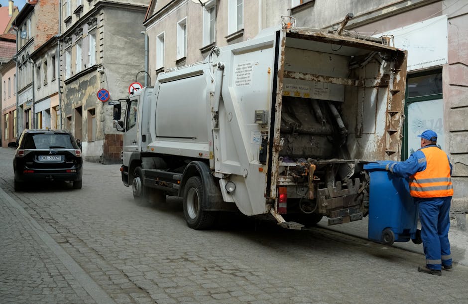 A top-down view of a grey metal trash bin filled with crumpled white paper, situated on a brown carpeted floor. Two additional crumpled sheets of paper are placed on the floor outside the bin. The sce