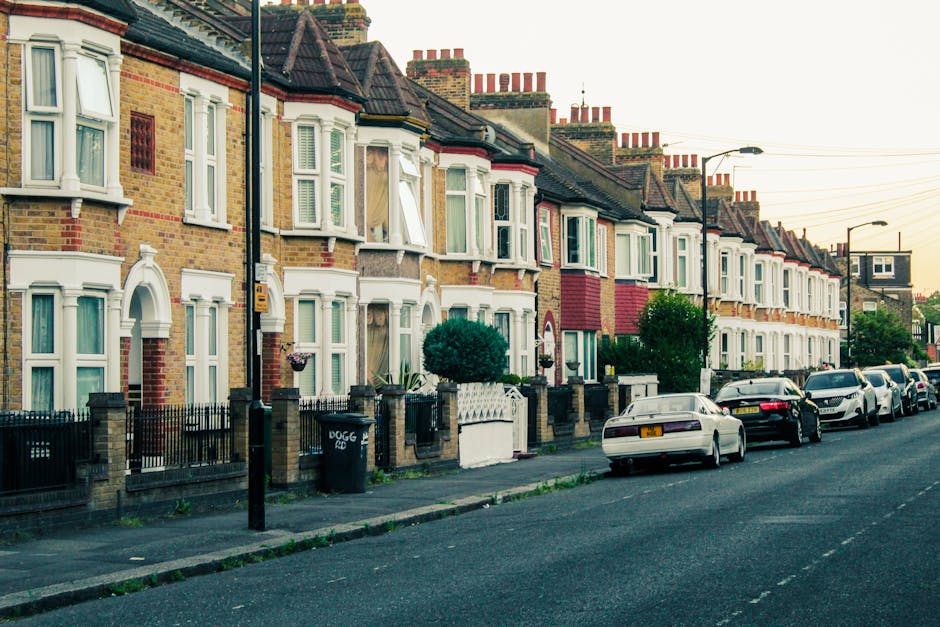 A row of Victorian-style terraced houses with brick facades, white-painted bay windows, and decorative trim along Wimbledon Hill Road in Merton. The houses feature pitched roofs with chimney stacks and are set behind small front gardens bordered by low brick walls and metal fencing. The pavement in front is clean, with parked cars lining the street, including a black sedan, a silver vehicle, and other contemporary cars. Soft evening light illuminates the street, highlighting the tidy appearance of the neighborhood, which benefits from professional domestic cleaning and surface cleaning services provided by Merton Cleaners to maintain its pristine condition.